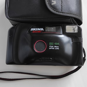 Op de afbeelding: Een zwarte en zilveren 35mm filmcamera met een rode lensring. De camera heeft een zwarte band en een zwarte tas met het woord "SKINA" erop gedrukt. De camera heeft de tekst "SK-106 F5.6 35mm JAPAN LENS" erop gedrukt.