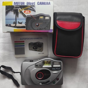 Puede incluir: Una c&aacute;mara de pel&iacute;cula de 35 mm plateada con una correa negra, una lente roja y las palabras "Protax" y "DX" impresas en la parte delantera. La c&aacute;mara est&aacute; sobre una superficie blanca.