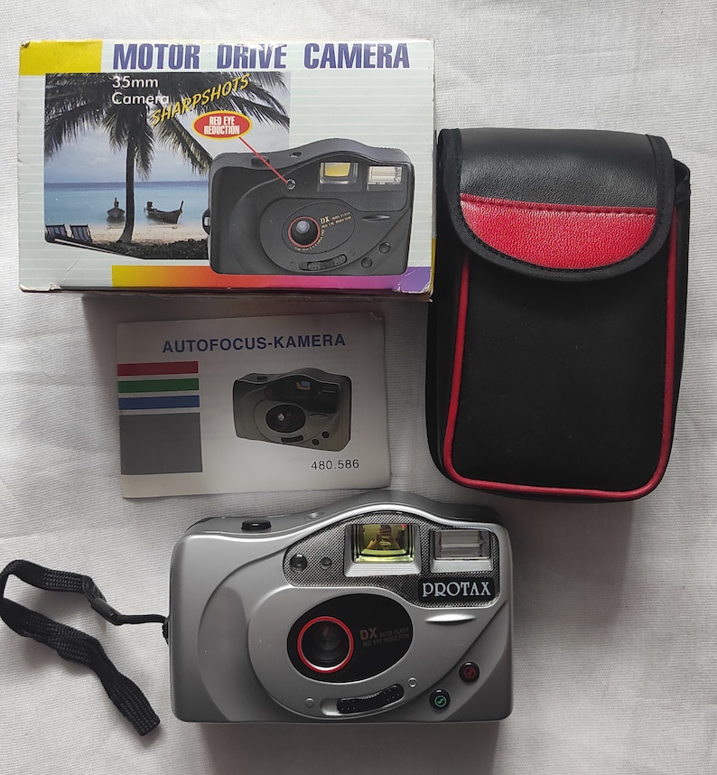 Puede incluir: Una c&aacute;mara de pel&iacute;cula de 35 mm plateada con una correa negra, una lente roja y las palabras "Protax" y "DX" impresas en la parte delantera. La c&aacute;mara est&aacute; sobre una superficie blanca.