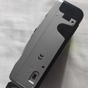 Puede incluir: Una c&aacute;mara compacta plateada y negra con una correa negra. La c&aacute;mara tiene una marca CE y un peque&ntilde;o bot&oacute;n negro.