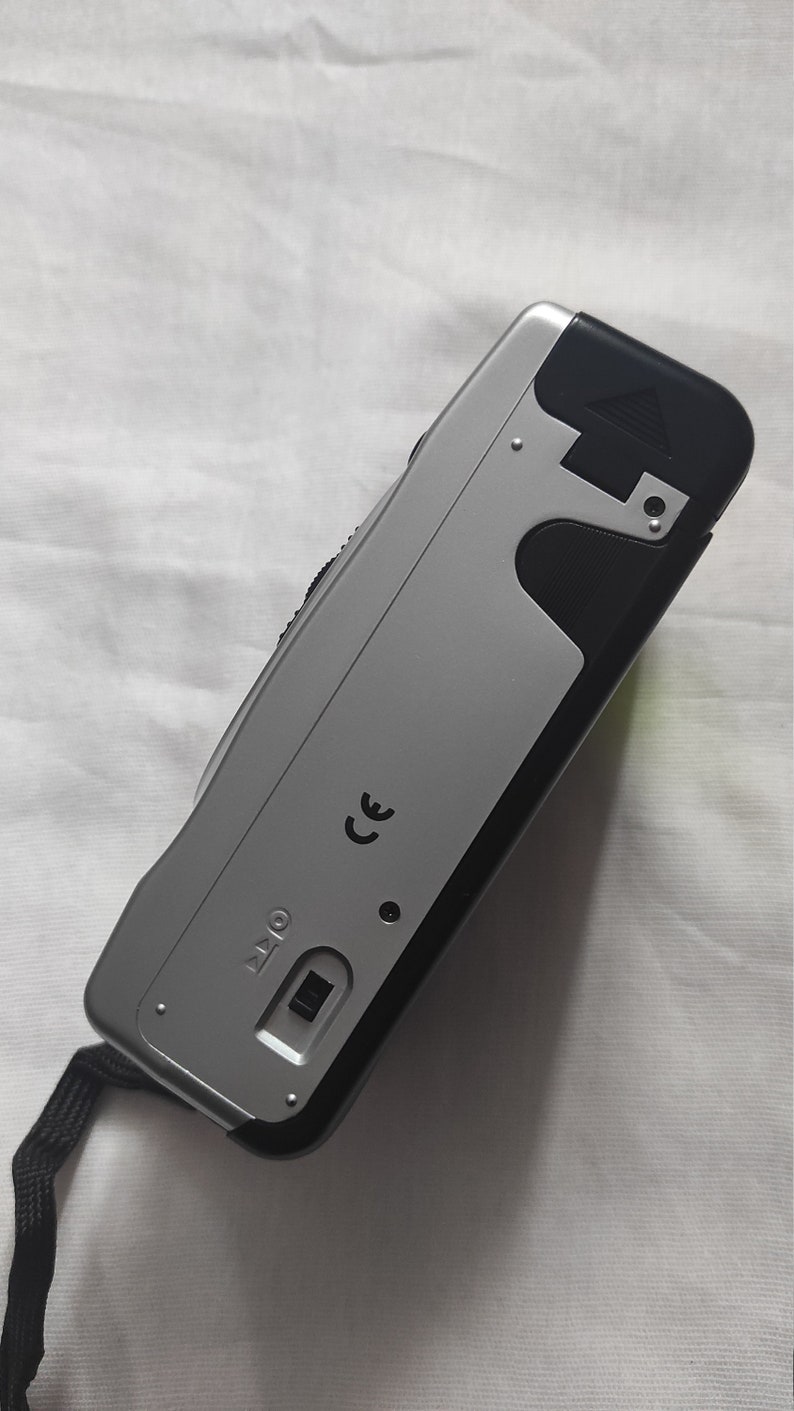 Puede incluir: Una c&aacute;mara compacta plateada y negra con una correa negra. La c&aacute;mara tiene una marca CE y un peque&ntilde;o bot&oacute;n negro.