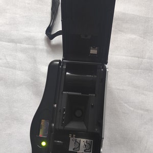 Puede incluir: Una c&aacute;mara de pel&iacute;cula negra y plateada con la parte posterior abierta, que revela el casete de pel&iacute;cula y una luz verde. La c&aacute;mara tiene una correa negra.