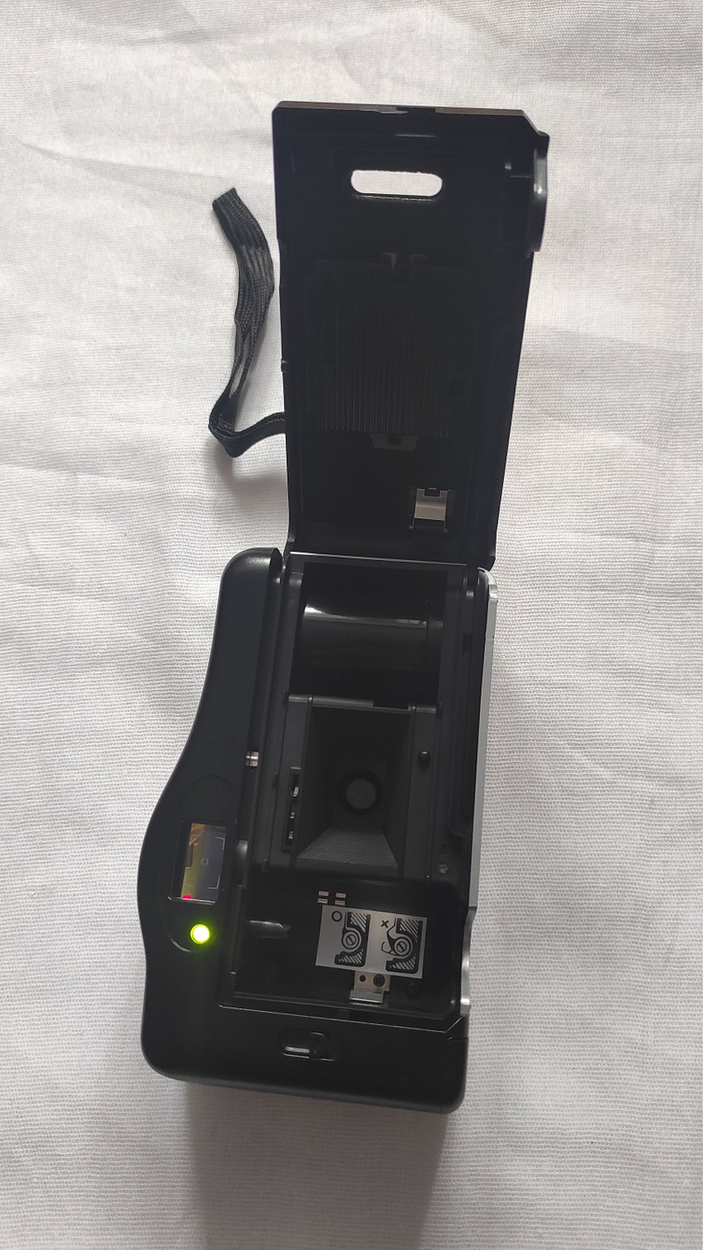 Puede incluir: Una c&aacute;mara de pel&iacute;cula negra y plateada con la parte posterior abierta, que revela el casete de pel&iacute;cula y una luz verde. La c&aacute;mara tiene una correa negra.