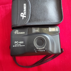 Puede incluir: Cámara de película de 35 mm Premier PC-660 negra con flash incorporado y reducción de ojos rojos. La cámara tiene una funda de cuero negra con el logotipo de Premier.