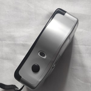 Puede incluir: Una c&aacute;mara de pel&iacute;cula compacta de color plateado y negro con una correa negra. La c&aacute;mara tiene una lente en la parte delantera y un bot&oacute;n en la parte superior.