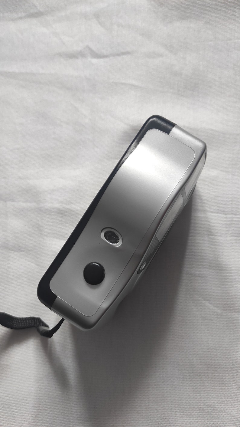 Puede incluir: Una c&aacute;mara de pel&iacute;cula compacta de color plateado y negro con una correa negra. La c&aacute;mara tiene una lente en la parte delantera y un bot&oacute;n en la parte superior.