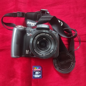 May include: A black Canon PowerShot S5 IS digital camera with a zoom lens and a black strap. The camera has an 8.0 megapixel resolution. A blue 32GB Transcend SD card and lens cap are also visible.