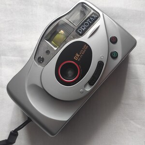 Puede incluir: Una c&aacute;mara compacta Protax DX plateada con una correa negra. La c&aacute;mara tiene un anillo rojo alrededor del objetivo y dice "DX Auto Flash Red Eye Reduction" en la parte delantera.