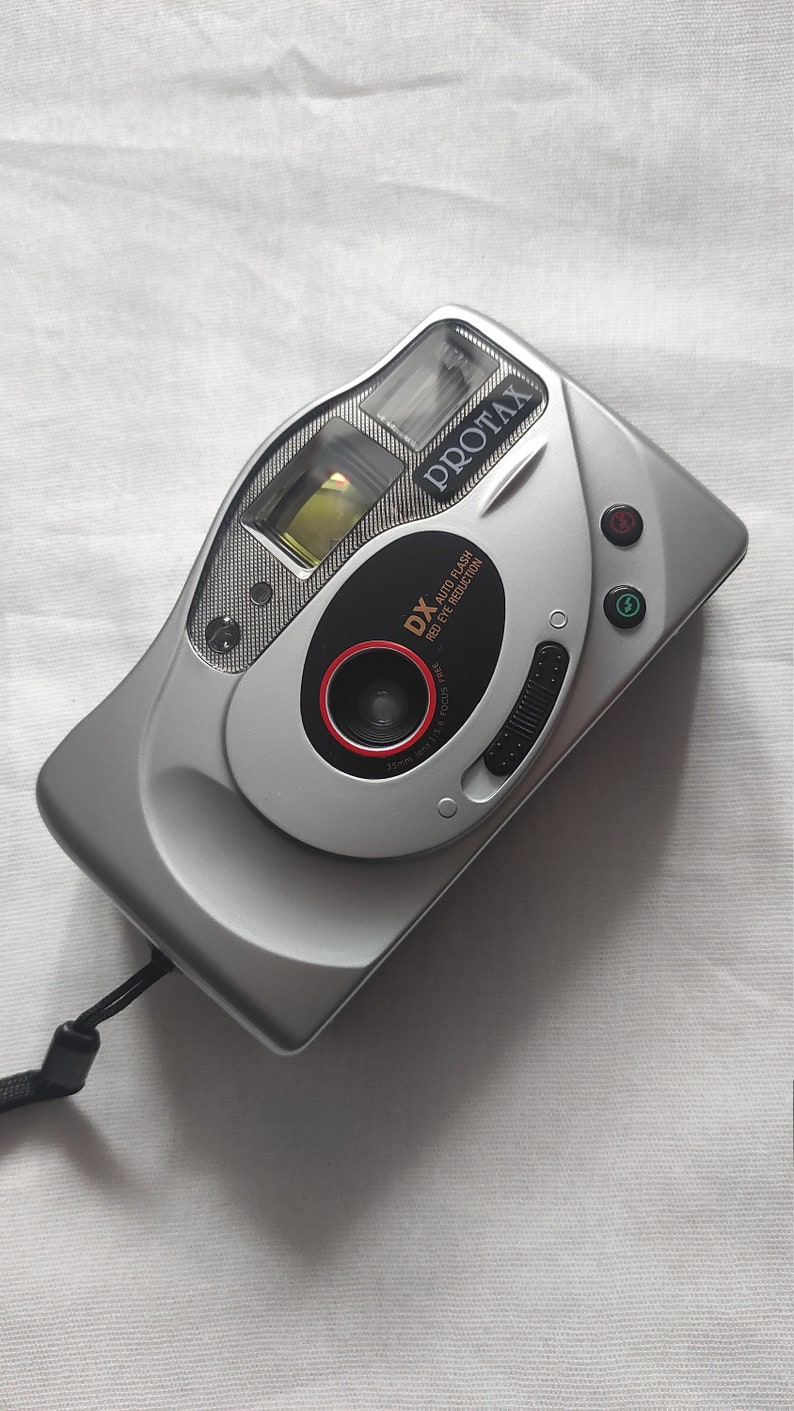Puede incluir: Una c&aacute;mara compacta Protax DX plateada con una correa negra. La c&aacute;mara tiene un anillo rojo alrededor del objetivo y dice "DX Auto Flash Red Eye Reduction" en la parte delantera.