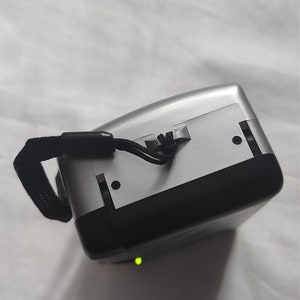 Puede incluir: Una c&aacute;mara digital plateada y negra con una correa negra. La c&aacute;mara tiene una luz verde en la parte inferior.