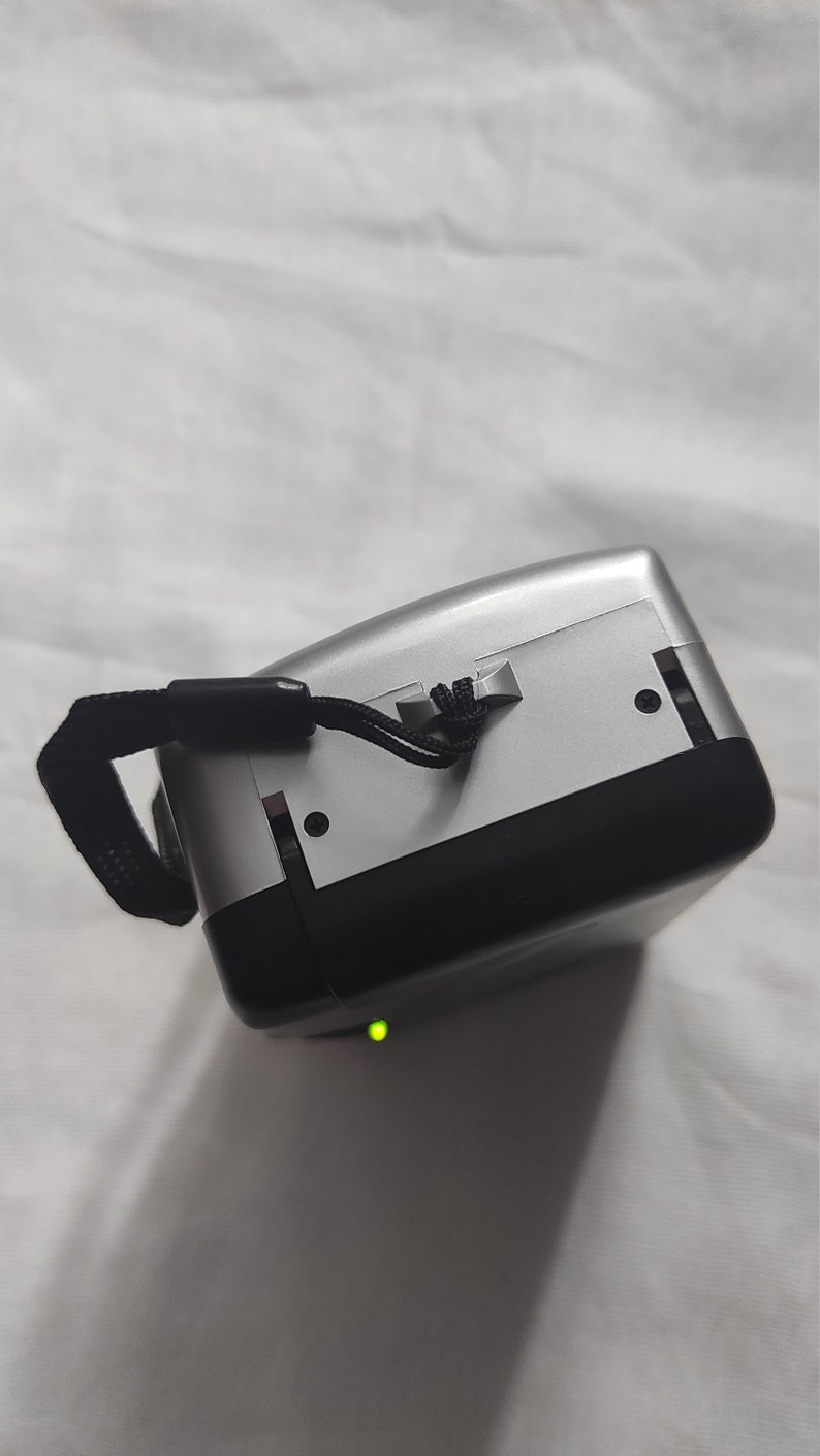 Puede incluir: Una c&aacute;mara digital plateada y negra con una correa negra. La c&aacute;mara tiene una luz verde en la parte inferior.