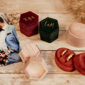 Könnte beinhalten: Sechs Samt-Ringboxen in verschiedenen Farben und Formen. Die Boxen sind mit Initialen in goldener Schrift personalisiert. Die Boxen befinden sich auf einer Holzoberfläche.