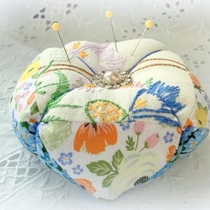 May include: A white fabric pincushion with a floral pattern and a pearl-like button in the center. Four yellow pins are stuck into the pincushion.