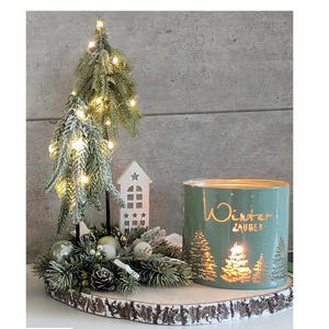 Könnte beinhalten: Winterliche Dekoration mit einem kleinen, beleuchteten künstlichen Weihnachtsbaum, einem grünen Kerzenhalter mit der Aufschrift "Winter Zauber" und dekorativen Elementen auf einem Holzsockel. Die Anordnung umfasst Tannenzapfen und Ornamente.