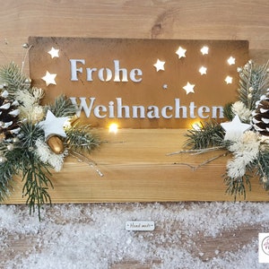 Pode incluir: Uma placa de madeira com as palavras "Frohe Weihnachten" em letras brancas sobre um fundo marrom rústico. A placa é decorada com estrelas brancas e luzes. A placa é cercada por ramos de pinheiro, bagas brancas e pinhas.