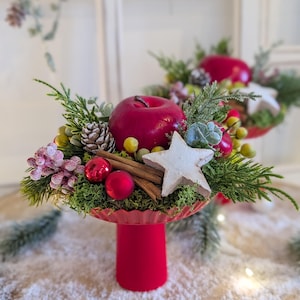 Könnte beinhalten: Eine festliche Tischdekoration mit einem roten Apfel, Zimtstangen und einem sternförmigen Ornament. Das Arrangement umfasst Grün, rote Kugeln und einen Tannenzapfen, alles in einer roten Schale auf einem Sockel. Zwei weitere Kugeln befinden sich im Vordergrund.
