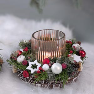 Könnte beinhalten: Weihnachtlicher Tischschmuck mit einem transparenten Glas-Kerzenhalter, umgeben von einem Kranz aus Grün, roten und weißen Kugeln, Beeren und weißen Sternen. Der Kranz steht auf einem Holzsockel.