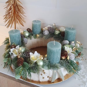 Può includere: Una ghirlanda dell'Avvento verde acqua con quattro candele verde acqua, un albero a stella dorato e decorazioni floreali bianche e verdi. La ghirlanda è decorata con pigne, bacche e piccole luci bianche.
