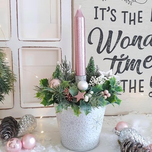Pode incluir: Um vaso de metal branco com um design de floco de neve cheio de um arranjo festivo de vegetação, enfeites rosa e prateados e uma vela rosa com glitter. O vaso está sobre uma superfície branca com o texto "It's the most wonderful time of the year" visível no fundo.
