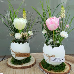 Può includere: Due gusci d'uovo bianchi con un arrangiamento di fiori di tulipano rosa e bianco all'interno di ciascuno. I gusci d'uovo sono appoggiati su una fetta di legno con muschio e un piccolo cartellino marrone che dice "Frohliche Ostergrüße" in tedesco.