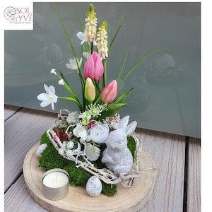 May include: A decorative Easter centerpiece on a wooden base. It features a gray bunny figurine, faux flowers including pink tulips, white blossoms, and green foliage. A small candle and a decorative egg are also included.