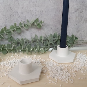 Könnte beinhalten: Zwei weiße Beton-Kerzenhalter mit sechseckigen Sockeln. Der Kerzenhalter rechts hat eine dunkelblaue Kerze darin.