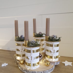 May include: A white and gold advent calendar made from repurposed cans with numbers 1, 2, and 3. The cans are decorated with gold ribbon and white twine. The candles are brown and are placed on top of the cans. The advent calendar is sitting on a wooden slice.