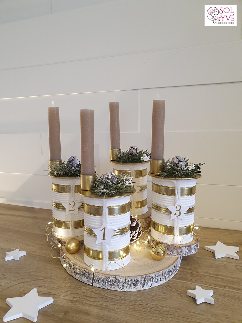 May include: A white and gold advent calendar made from repurposed cans with numbers 1, 2, and 3. The cans are decorated with gold ribbon and white twine. The candles are brown and are placed on top of the cans. The advent calendar is sitting on a wooden slice.