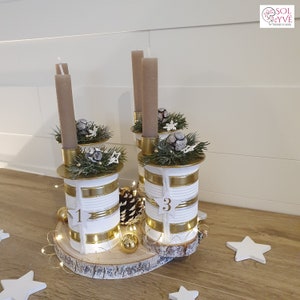 May include: A white and gold Christmas advent candle holder made from repurposed cans. The holder has four candles and is decorated with pine branches, pinecones, and snowflakes. The numbers 1 and 3 are visible on the cans.