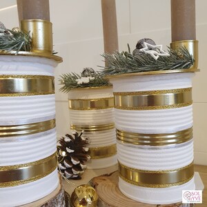 May include: Three white metal cans with gold and silver trim, decorated with pine branches and a star, used as candle holders. The cans are sitting on a wooden surface.
