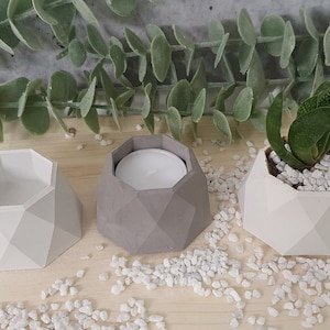 Peut inclure: Trois pots en béton géométriques, deux en blanc et un en gris. Le pot gris contient une bougie chauffe-plat blanche. Les pots blancs sont remplis de gravier blanc et l'un d'eux contient une petite plante succulente verte.
