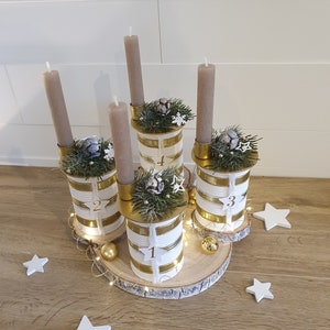 May include: A white and gold advent candle holder with four candles, decorated with greenery and white snowflakes. The holder is made from repurposed cans and sits on a wooden slice.