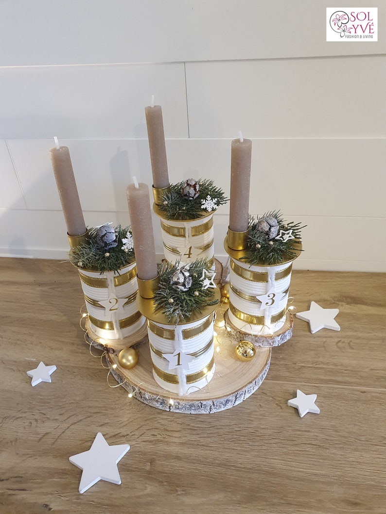 May include: A white and gold advent candle holder with four candles, decorated with greenery and white snowflakes. The holder is made from repurposed cans and sits on a wooden slice.