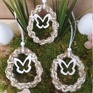 Può includere: Tre ghirlande fatte a mano in iuta e corda bianca con un ritaglio di farfalla bianca al centro. Le ghirlande sono appese a un nastro bianco e sono esposte su un letto di muschio verde. Il testo "Hand made" è visibile nell'angolo in basso a destra dell'immagine.