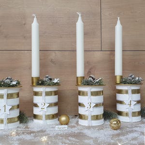 May include: Four white candlesticks with gold accents and numbers 1 through 4 on each. Each candlestick is decorated with a small sprig of greenery and gold ribbon.