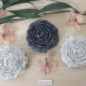 Puede incluir: Tres piezas de decoración de pared de flores de papel hechas a mano. Una es gris, otra es blanca y otra es de color melocotón claro. Las flores están dispuestas sobre una superficie de madera con hojas verdes.