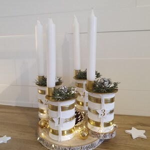 May include: A white and gold advent candle holder made from repurposed tin cans. The holder is decorated with twine, gold ribbon, and pine sprigs. The candles are white and the numbers 1, 2, 3, and 4 are visible on the cans.