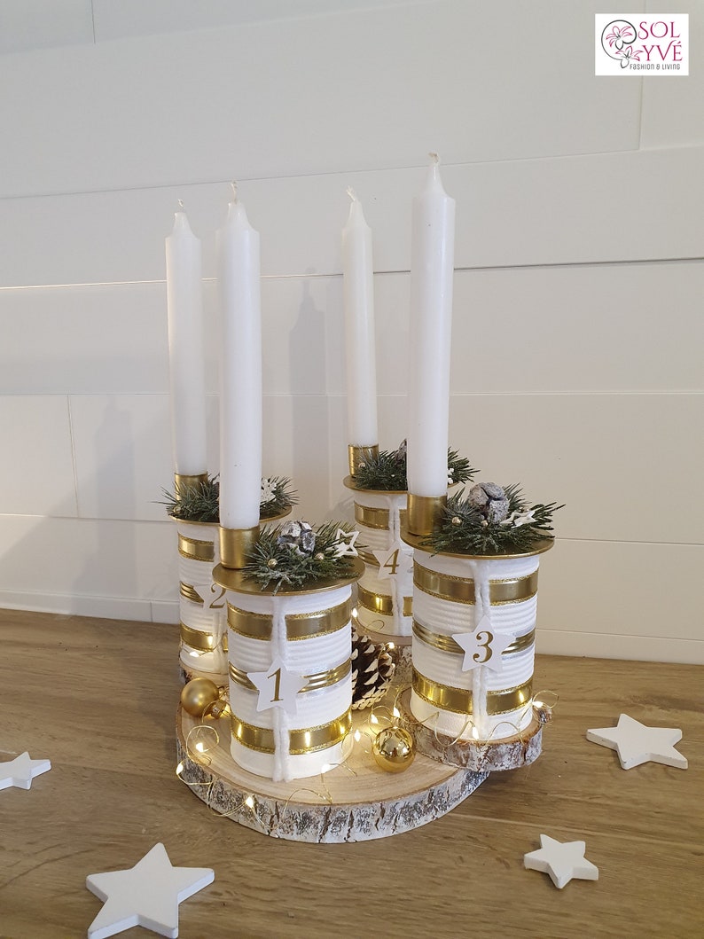 May include: A white and gold advent candle holder made from repurposed tin cans. The holder is decorated with twine, gold ribbon, and pine sprigs. The candles are white and the numbers 1, 2, 3, and 4 are visible on the cans.