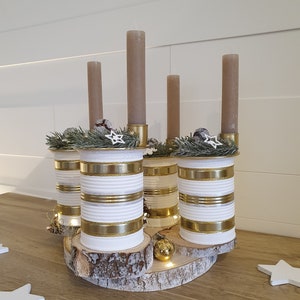 May include: A rustic Christmas centerpiece made from repurposed tin cans painted white with gold stripes. The cans are arranged in a circle on a wooden slice and topped with candles and greenery.