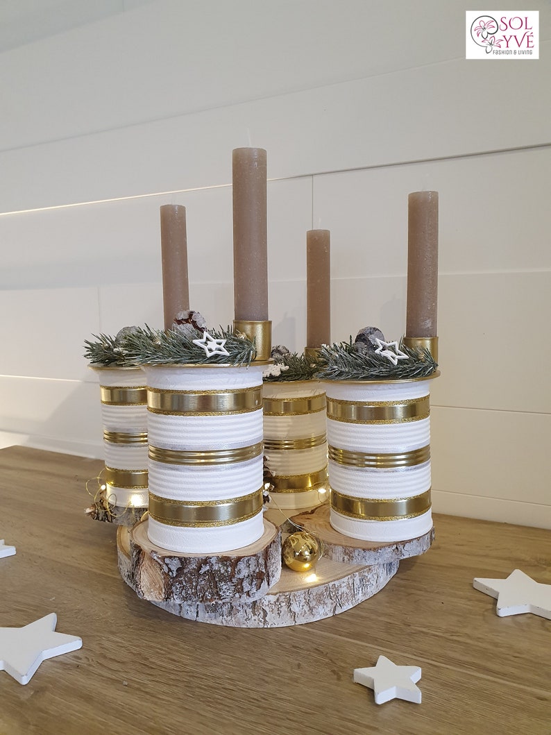 May include: A rustic Christmas centerpiece made from repurposed tin cans painted white with gold stripes. The cans are arranged in a circle on a wooden slice and topped with candles and greenery.