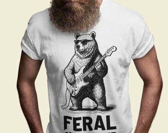 Può includere: T-shirt bianca con un'illustrazione in bianco e nero di un orso con occhiali da sole che suona una chitarra elettrica. Il testo "FERAL OLD & LOUD" è stampato sotto la grafica dell'orso. La maglietta ha maniche corte.