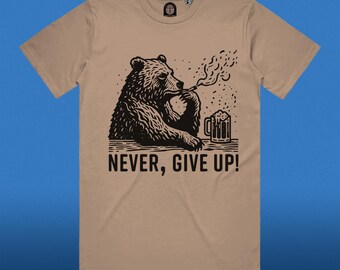 Può includere: Una maglietta color beige con un'illustrazione in bianco e nero di un orso che fuma una sigaretta e tiene una birra. Il testo "NEVER, GIVE UP!" è stampato sotto l'orso.
