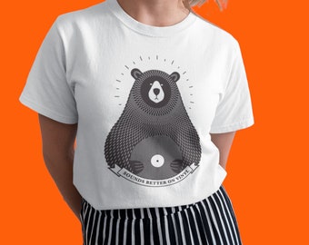 Può includere: Maglietta bianca con un'illustrazione in bianco e nero di un orso che tiene un disco in vinile. Il testo "Sounds better on vinyl" è stampato sotto l'orso.