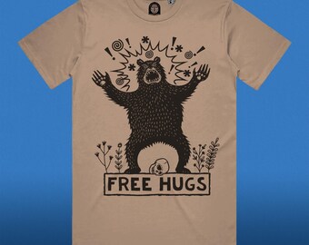 Può includere: Una maglietta color beige con un'illustrazione nera di un orso con le braccia alzate e il testo "FREE HUGS" sotto l'orso.