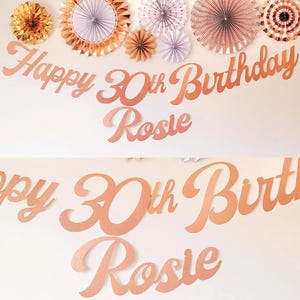 May include: Rose gold Happy 30th Birthday banner with the name Rosie. The banner is made of cardboard and has a cursive font.