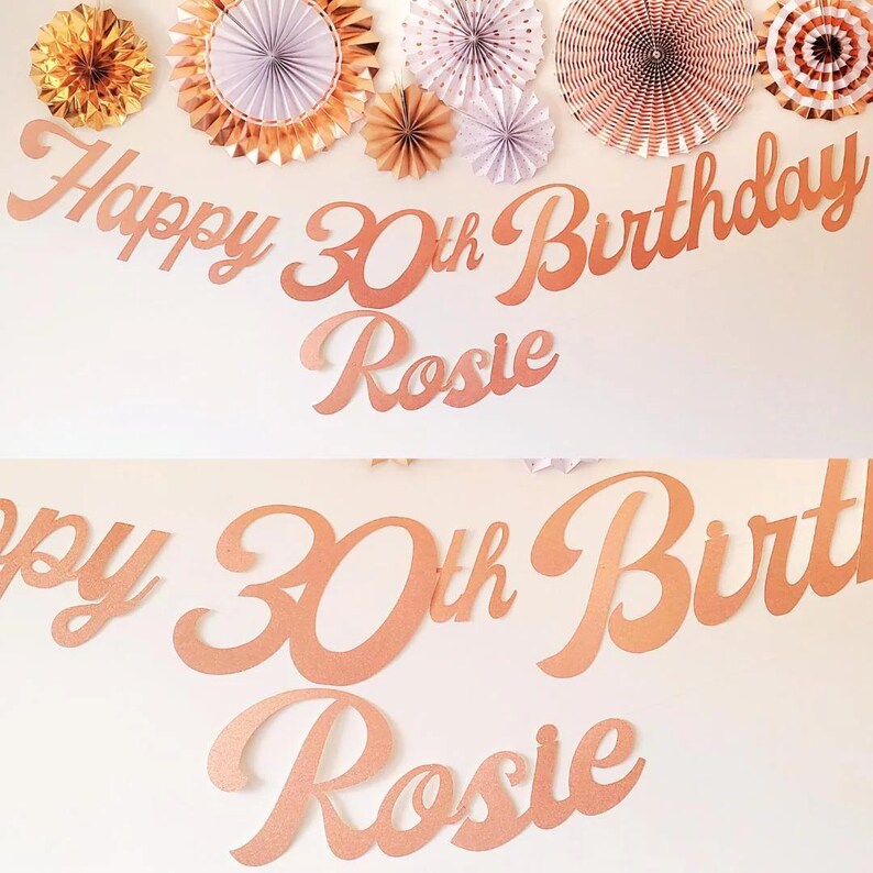 May include: Rose gold Happy 30th Birthday banner with the name Rosie. The banner is made of cardboard and has a cursive font.