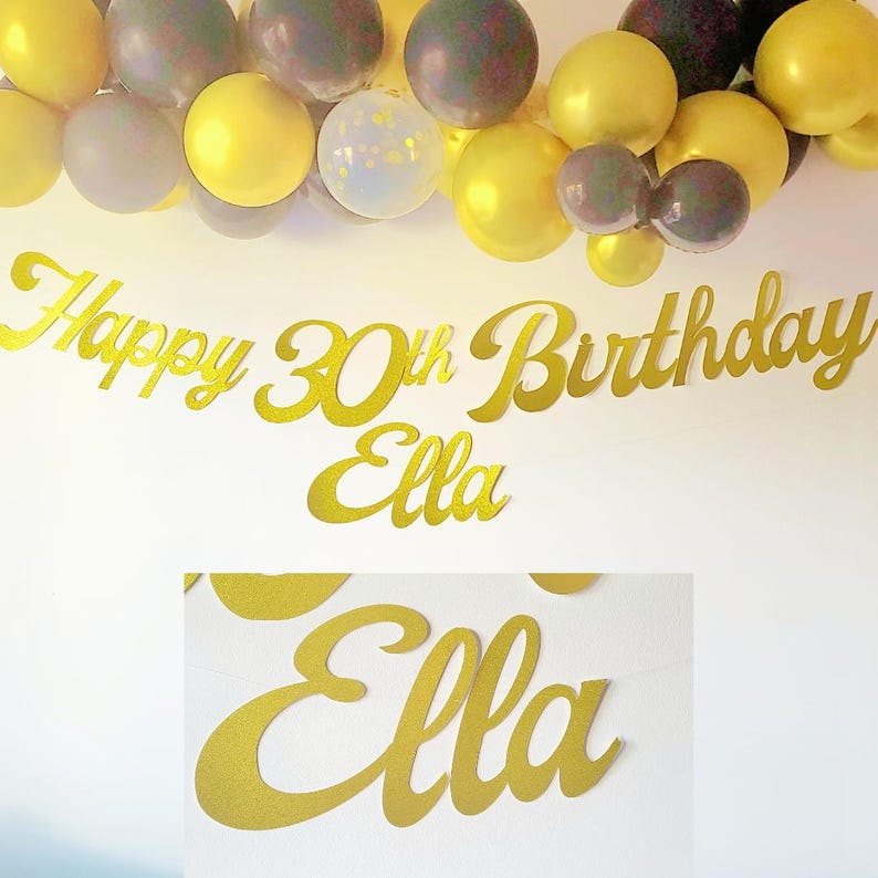 May include: Gold Happy 30th Birthday banner with the name Ella. The banner is decorated with gold and black balloons.
