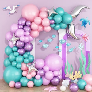 May include: A mermaid-themed balloon arch featuring pastel pink, purple and teal balloons. The arch is decorated with silver stars, fish and a silver mermaid tail. The background is a light purple wall.