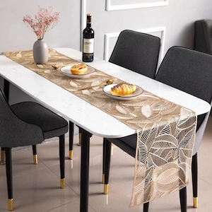 May include: A gold leaf patterned table runner on a white marble table with black legs. The runner is on a table set with two chairs and a bottle of wine.