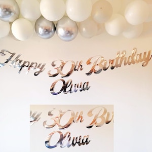 May include: Silver mirror-like Happy 30th Birthday Olivia banner. The banner is made of individual letters and numbers. The banner is hanging on a white wall with a silver and white balloon garland above it.